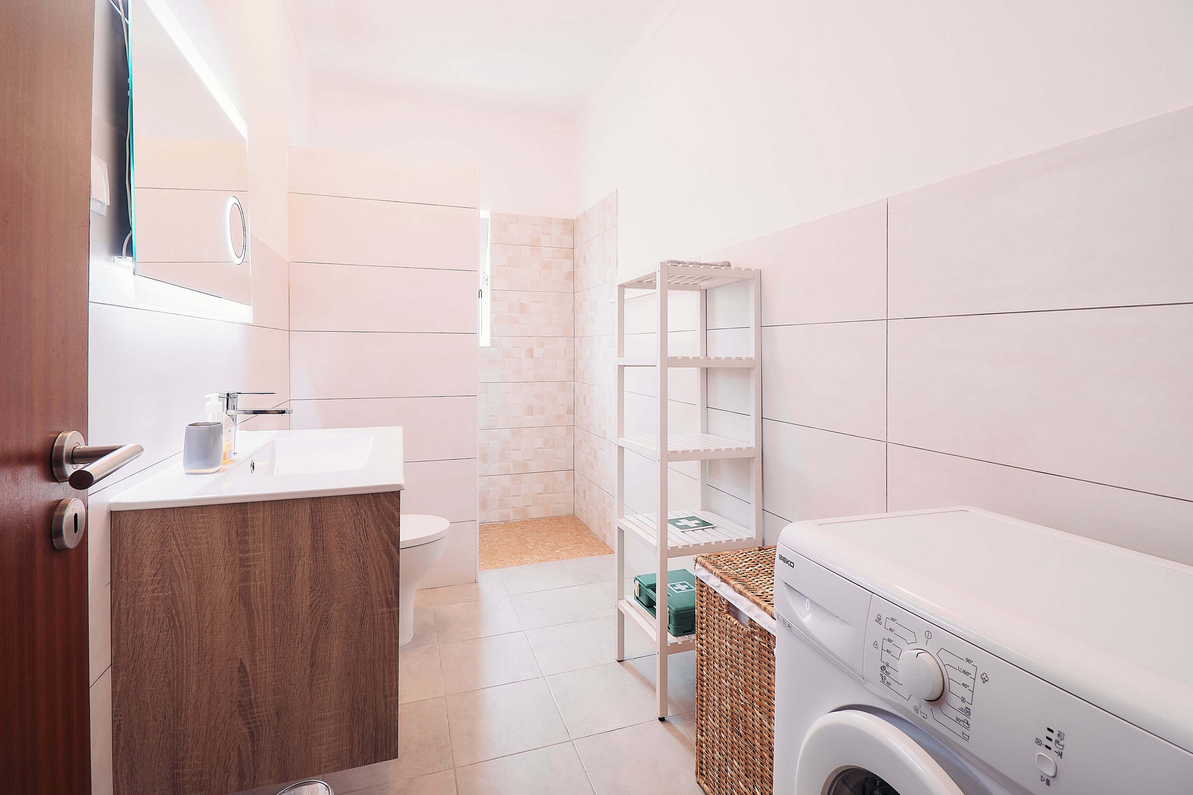 A compact bathroom with a wooden vanity, a toilet, a washing machine, and a shelf unit.