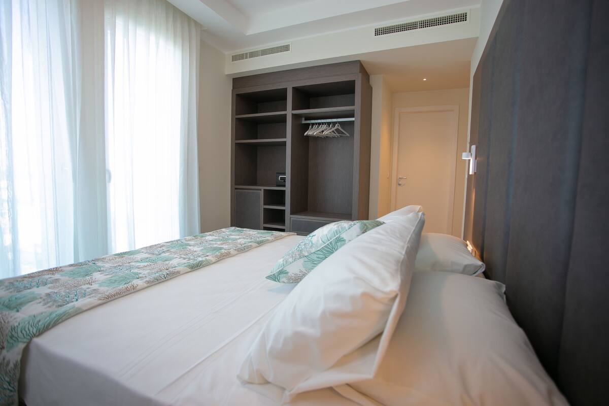 Un lit bien fait, recouvert d’une couvre-lit à motifs floraux, une armoire avec une tringle pour suspendre les vêtements, et une porte sur la droite.