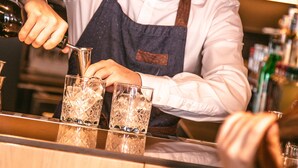 En bartender som står bakom bardisken och gör i ordning drinkar. Bakom honom finns hyllor fulla med flaskor och glas.