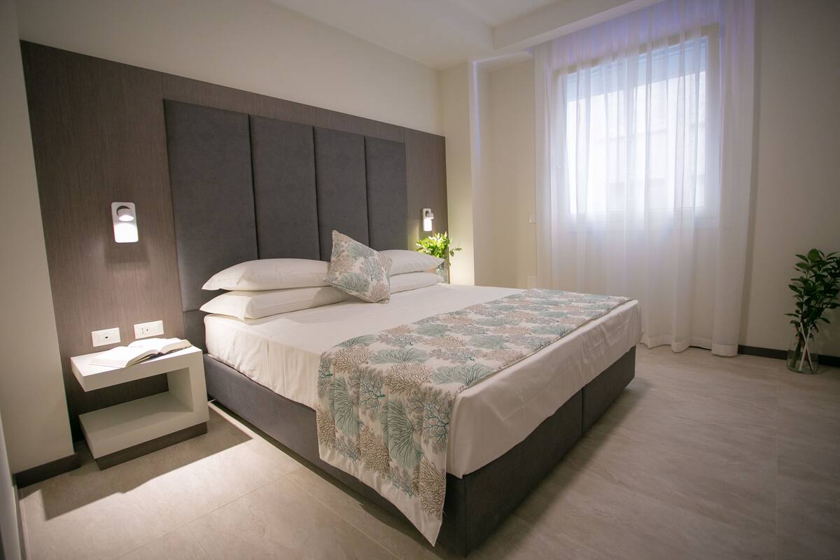 Une chambre moderne avec un grand lit, des tables de chevet et une fenêtre ornée de rideaux légers.