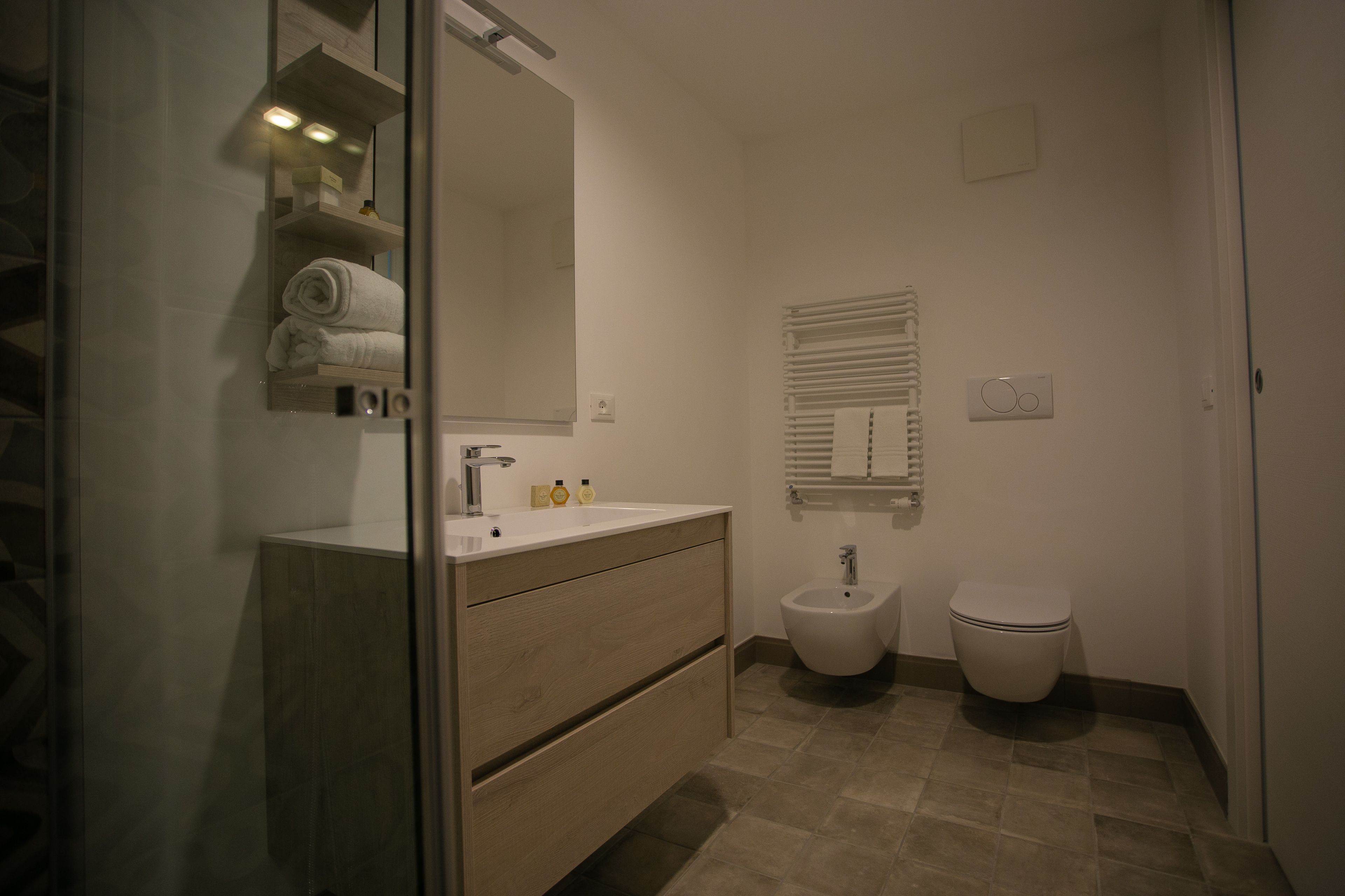 Een moderne badkamer met een glazen douchecabine, een houten wastafelmeubel met een spiegel, een bidet en een toilet.