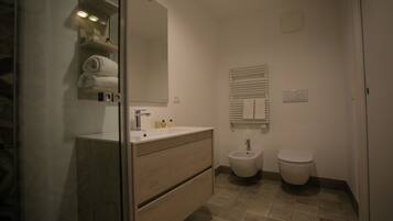 Een moderne badkamer met een glazen douchecabine, een houten wastafelmeubel met een spiegel, een bidet en een toilet.