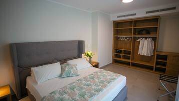 Een net opgemaakt bed met een bedsprei met patroon, een grijs gestoffeerd hoofdbord, en een nachtkastje met een lamp en een bloemenvaas.