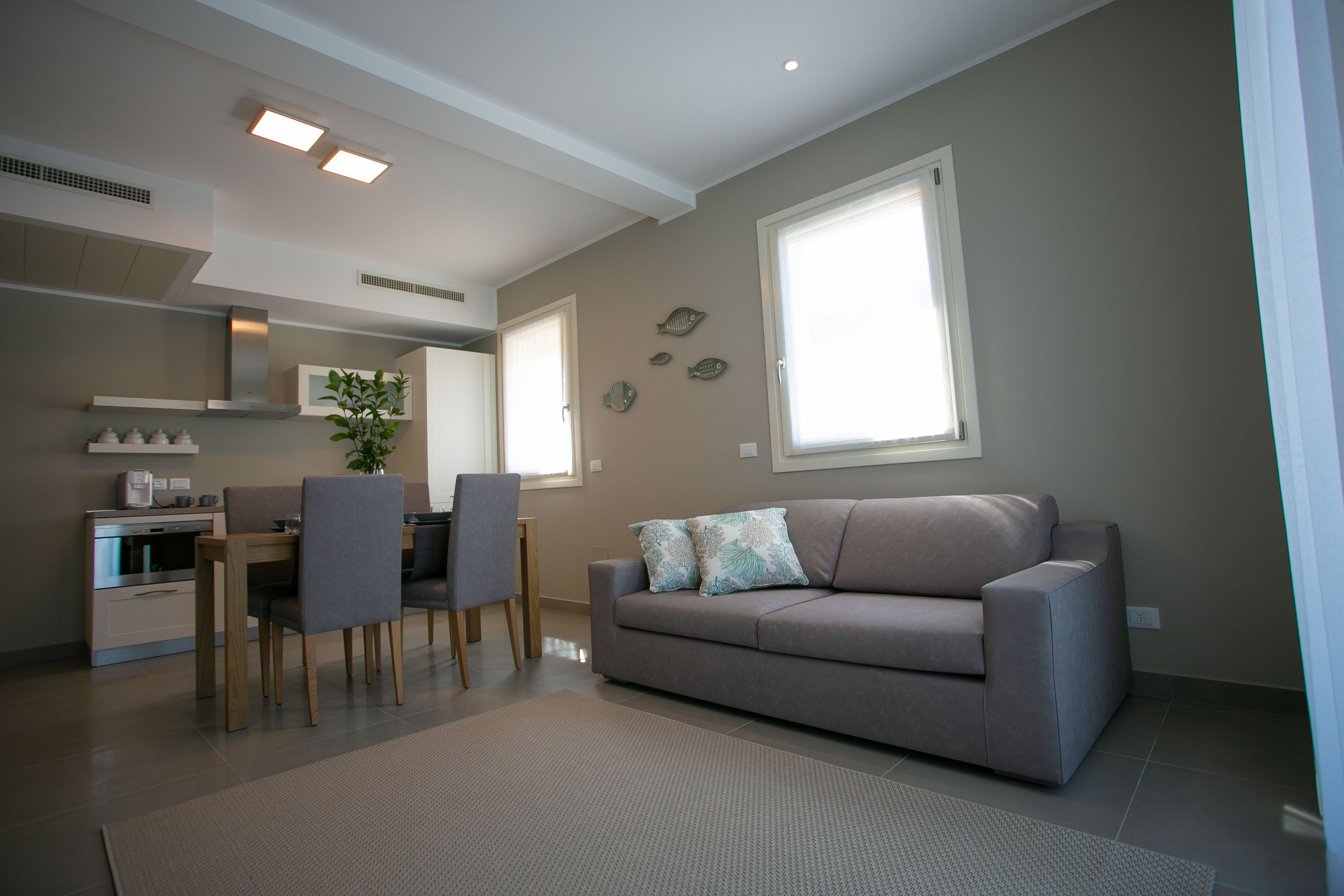 Un salon moderne avec un canapé gris, une table à manger avec des chaises et, au fond, un coin cuisine.