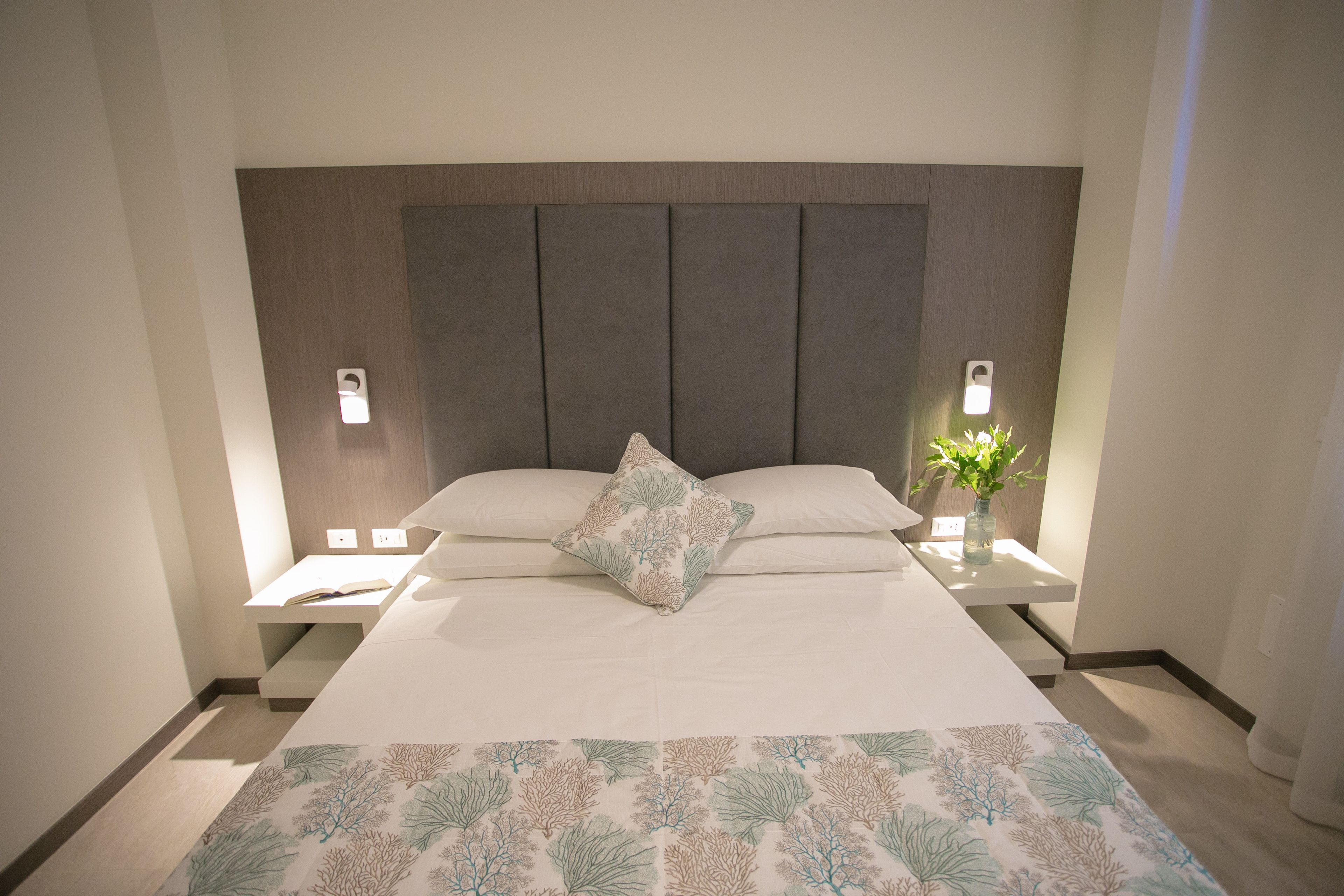 A neatly made bed with a patterned bedspread, two bedside tables with lamps, and a nightstand with a vase of flowers.
