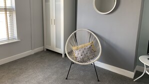 A room with a white wardrobe, a wicker chair with patterned cushions, a round mirror on the wall, and a small white table. - Buccleuch Apartment (Kettering)