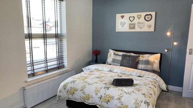 A bedroom with a bed, a radiator, a window with blinds, a wall with heart-shaped decorations, and a bedside lamp. - Buccleuch Apartment (Kettering)
