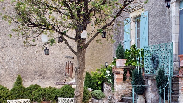 Un área de estar al aire libre con sillas de metal blanco y una mesa, rodeada de plantas en macetas y un árbol.