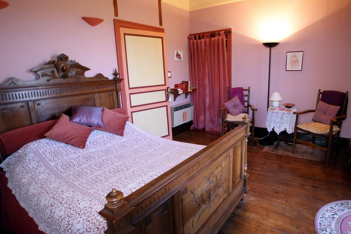 A bedroom with a wooden bed, a chair, a small table with a lamp, and a curtain.
