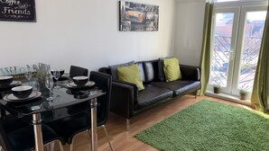 A modern living room with a dining area, featuring a black leather sofa, a glass dining table, and a cityscape view through a large window. - Eskdaill Place Apartment (Kettering)
