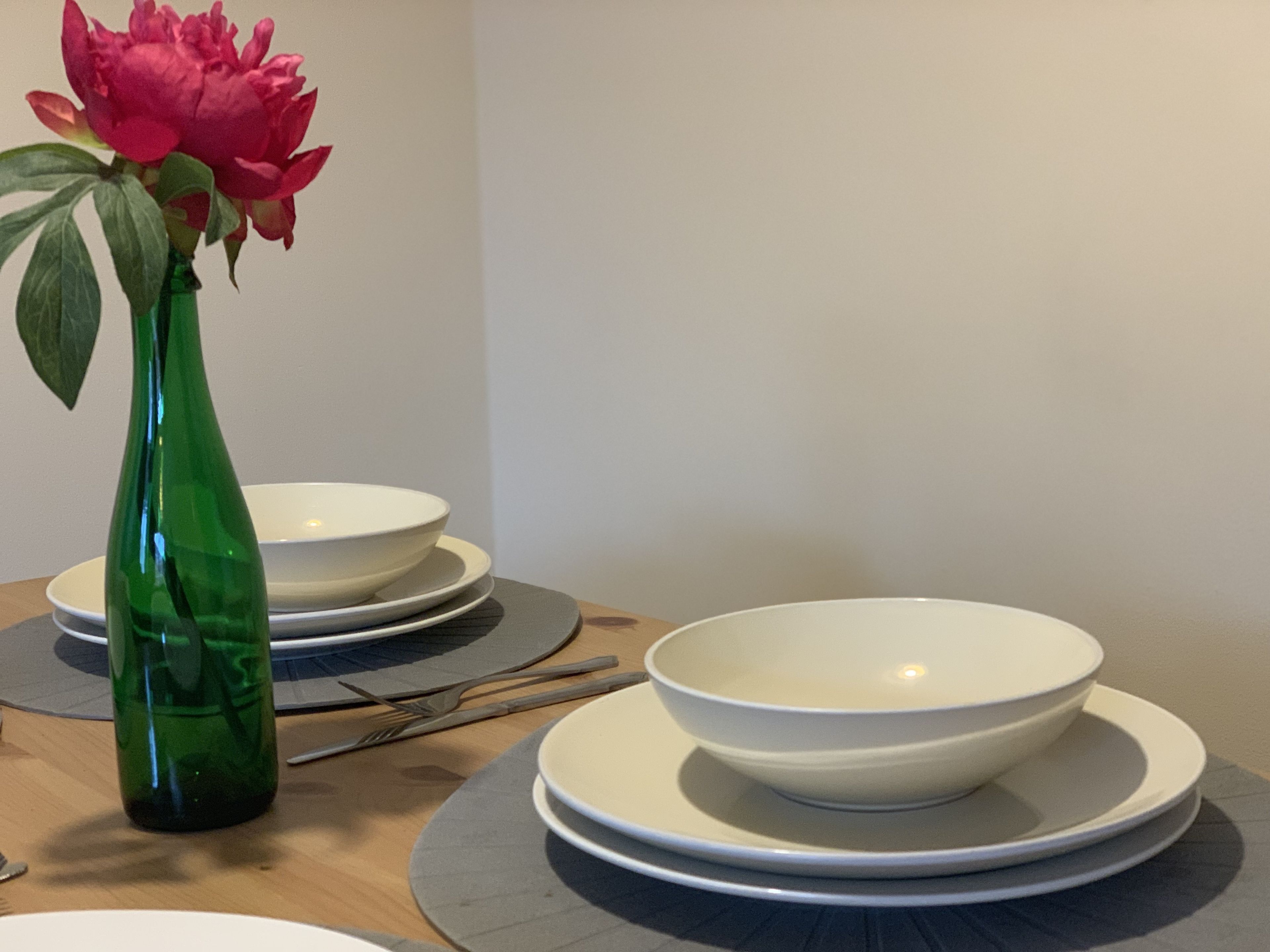Une table à manger ornée d’un vase vert contenant une fleur rose, de bols blancs et de couverts.