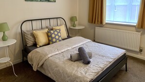 A bedroom with a bed, a green lamp, a white radiator, and a window with curtains. - Cosy Corner House (Kettering)