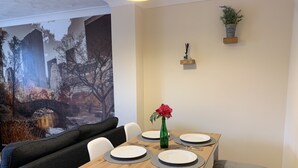 A dining area with a table set for four, a green vase with a flower, and a mural of a bridge and trees on the wall. - Cosy Corner House (Kettering)