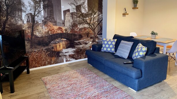 A living room with a blue sofa, patterned rug, and a cityscape mural on the wall. - Cosy Corner House (Kettering)
