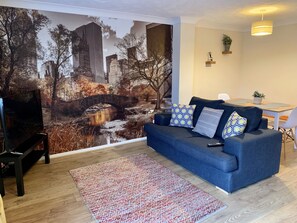 Un salon avec un canapé bleu, un tapis à motifs et une fresque représentant un paysage urbain sur le mur.
