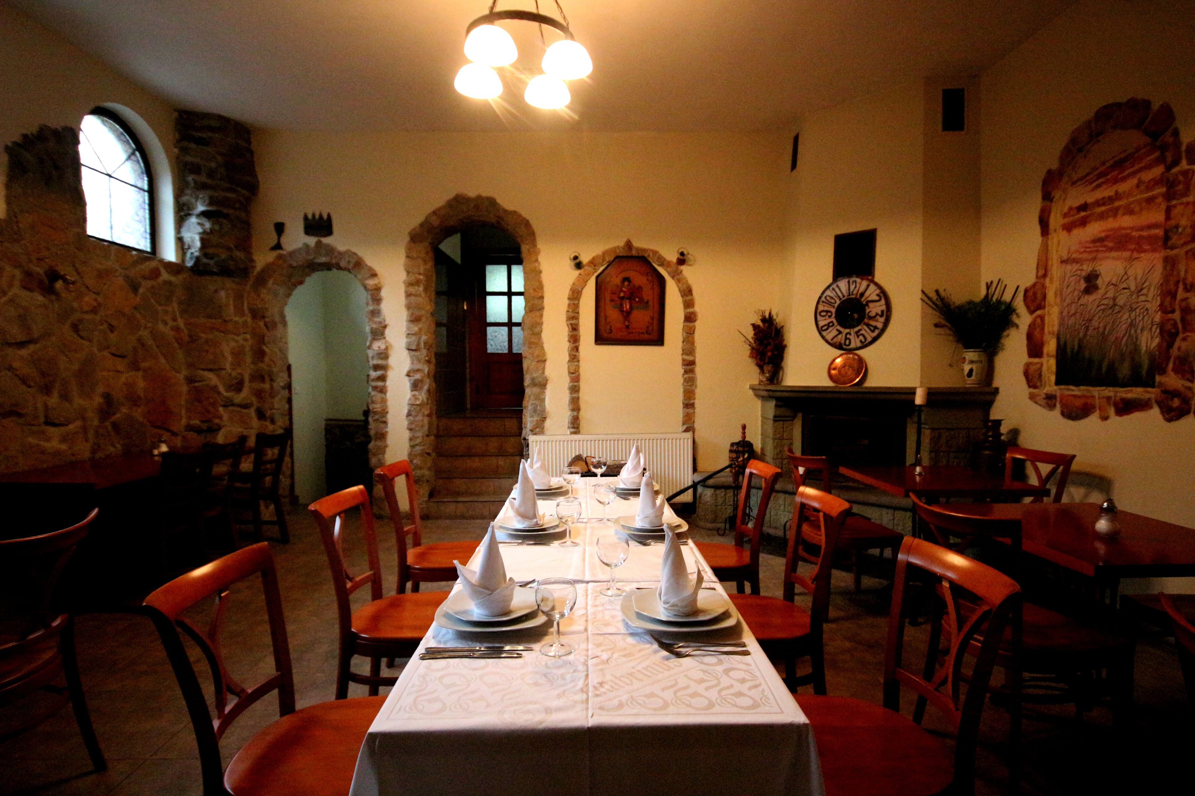 A dining room with a long table set for four, surrounded by wooden chairs. An arched doorway and a stone wall are visible in the background.
