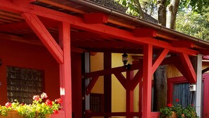 A covered outdoor seating area with red wooden beams, potted plants with red and yellow flowers, and a black dog on the ground. - Hotel Bor (Lhota)
