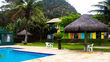 Uma piscina com deck de pedra, cercada por palmeiras e um bangalô com telhado de palha.