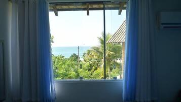 Quarto com uma grande janela que oferece vista para o oceano e vegetação exuberante.