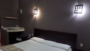 Una habitación de hotel con una cama, una mesita de noche, una lámpara de pared y un pequeño estante.