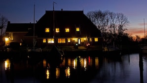 A waterfront building with multiple lit windows, a dock with boats, and a calm water body reflecting the lights.