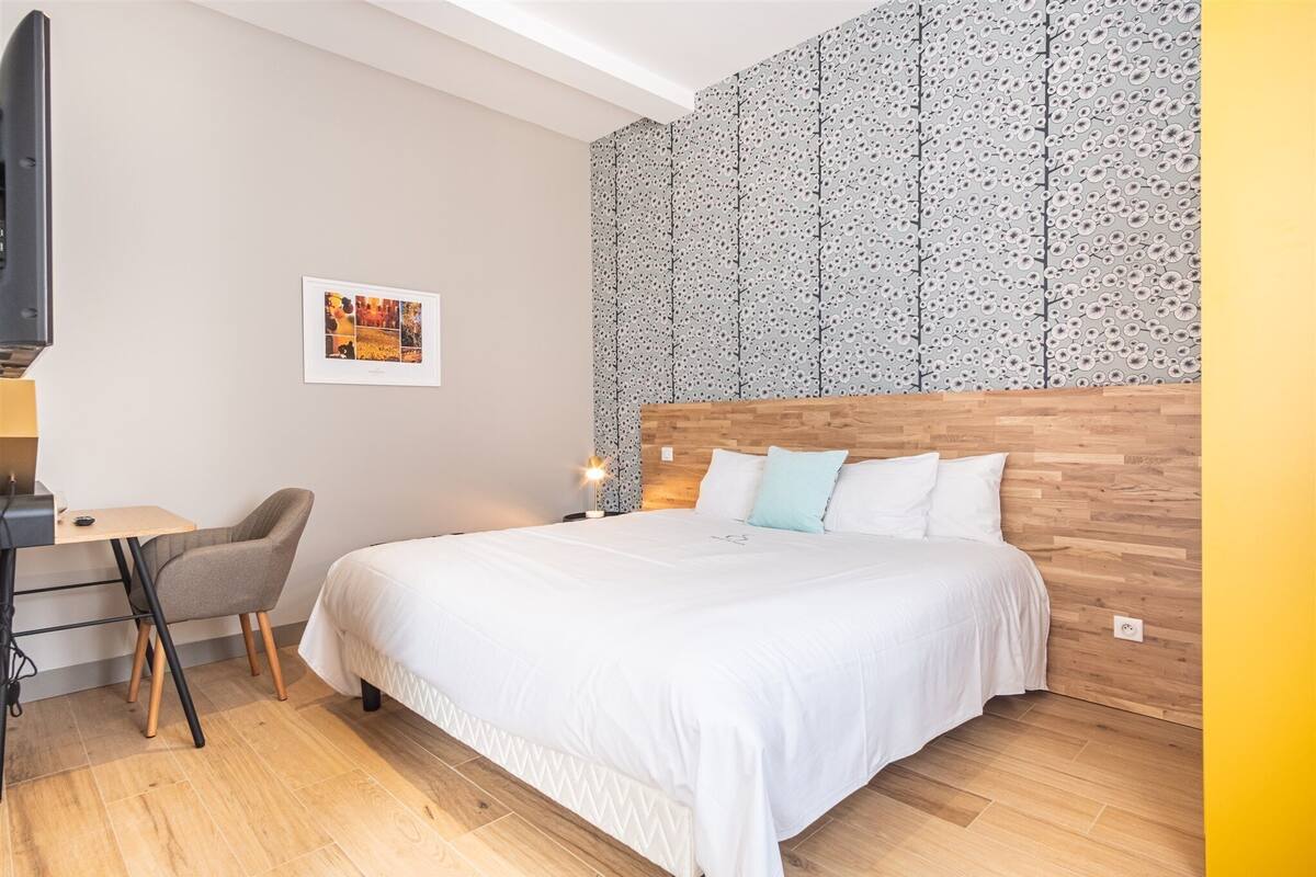 A modern hotel room with a wooden headboard, a bed with white bedding, a desk with a chair, a flat-screen TV, and a patterned wall.