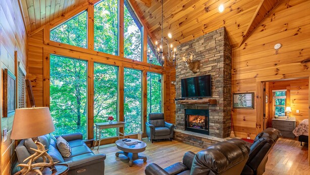 A cozy cabin interior with a stone fireplace, leather sofas, and large windows offering a view of the forest. - Cardinal's Nest- Along Cranberry Creek, great for tubing, kayaking (Elk Park)