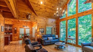 A log cabin interior with a fireplace, leather furniture, and large windows offering a view of the forest. - Cardinal's Nest- Along Cranberry Creek, great for tubing, kayaking (Elk Park)