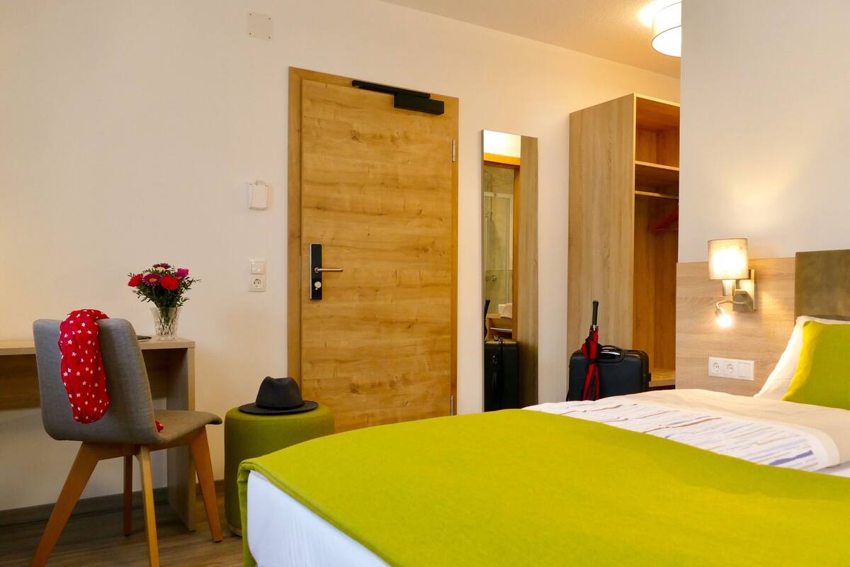 Una habitación de hotel con una cama, una silla, una mesita, un espejo y un armario.