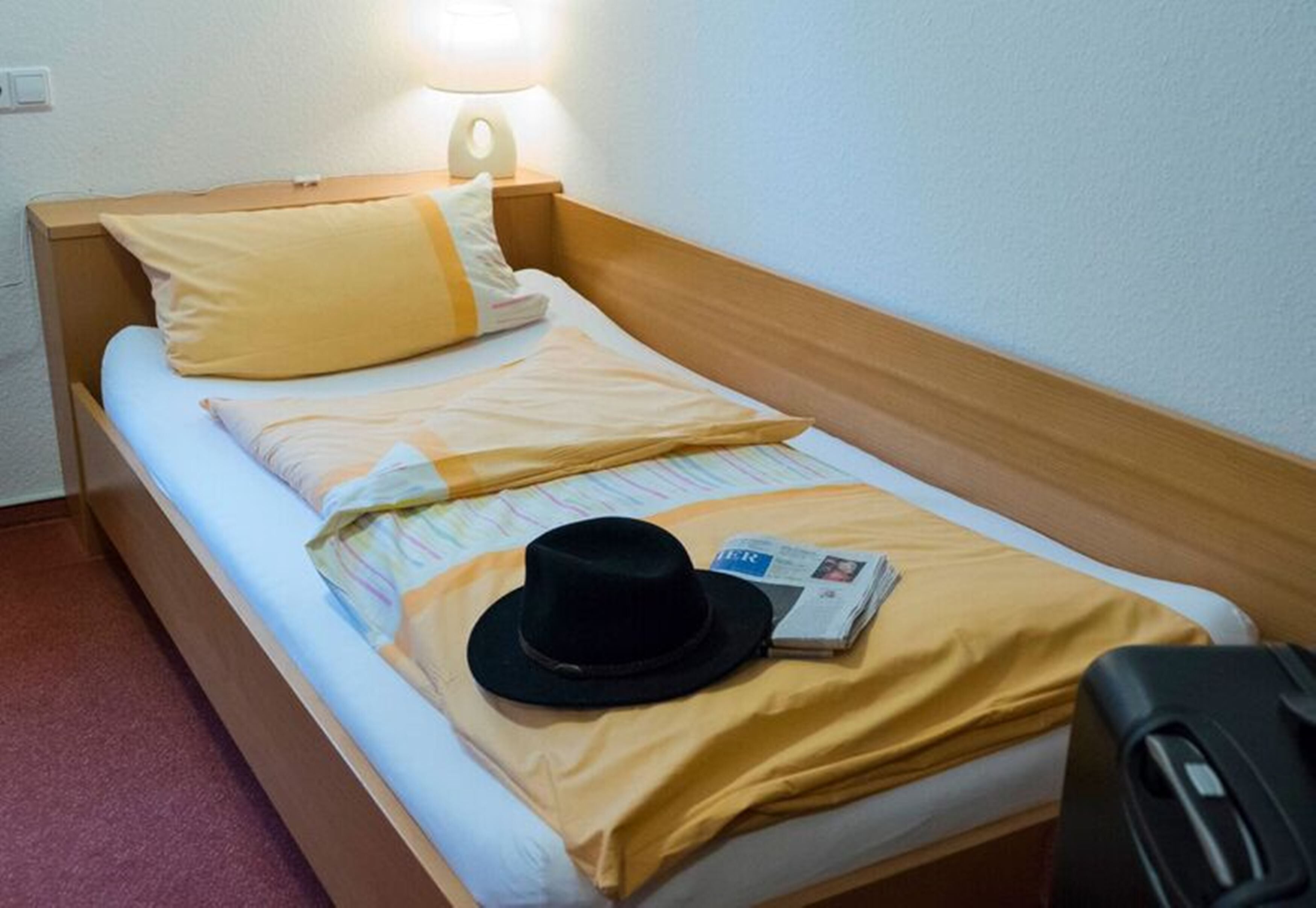 Una cama individual con cabecera de madera, un almohón amarillo, un sombrero negro y un periódico sobre la cama.