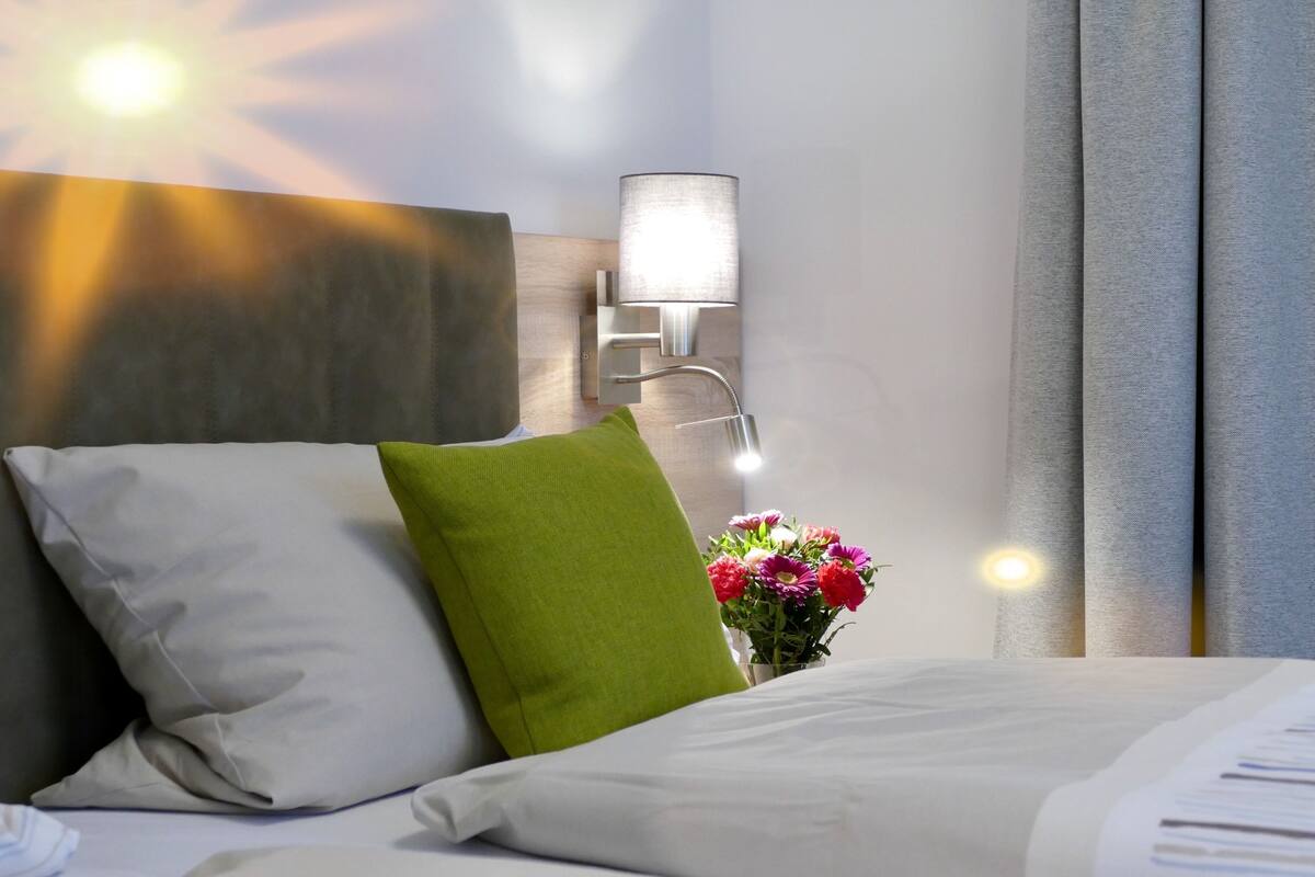 Una habitación de hotel con cama, almohadas, una mesita de noche con lámpara y un jarrón de flores.