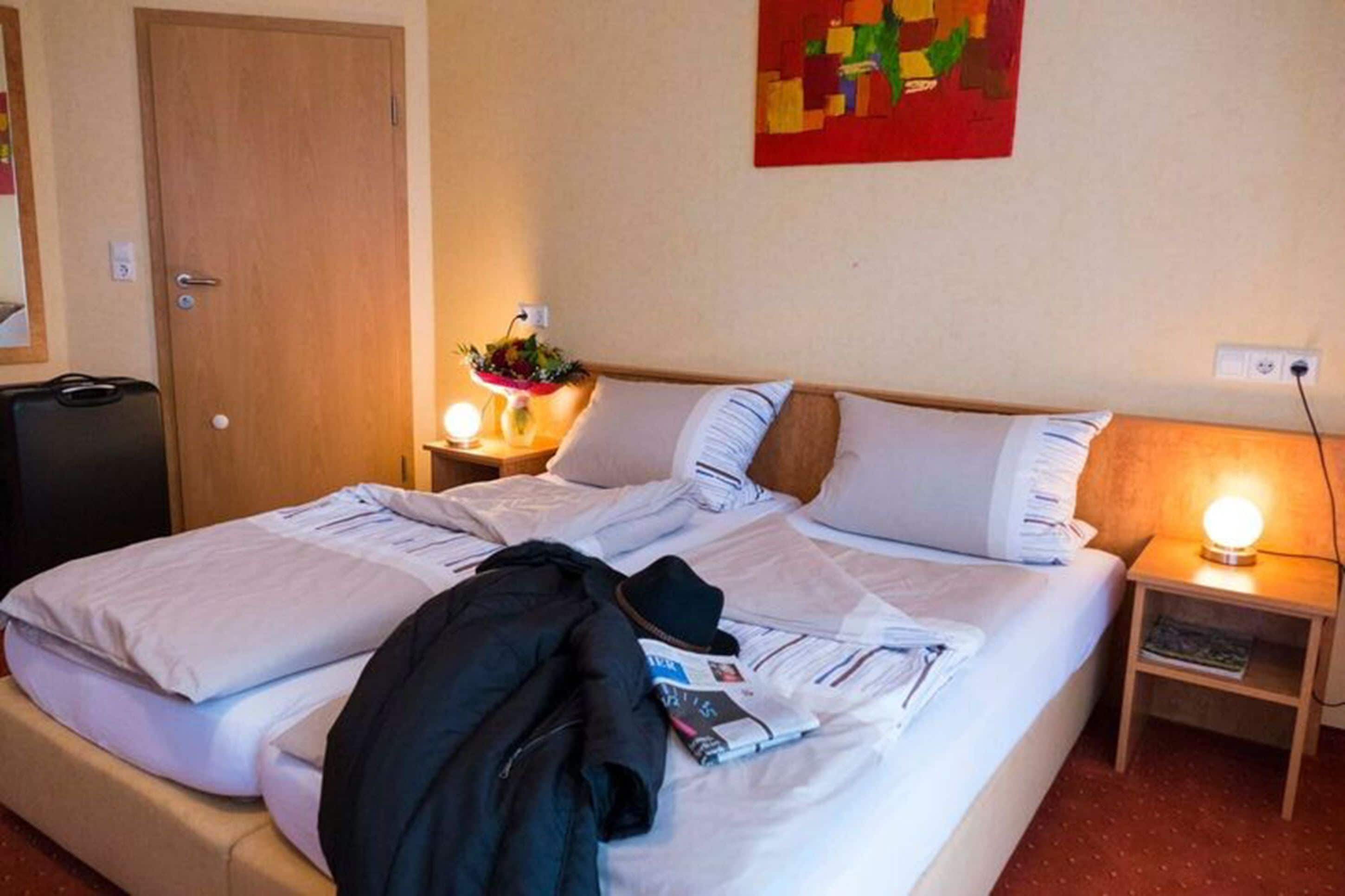A hotel room with a bed, a nightstand with a lamp, a small table with a flower vase, and a door.