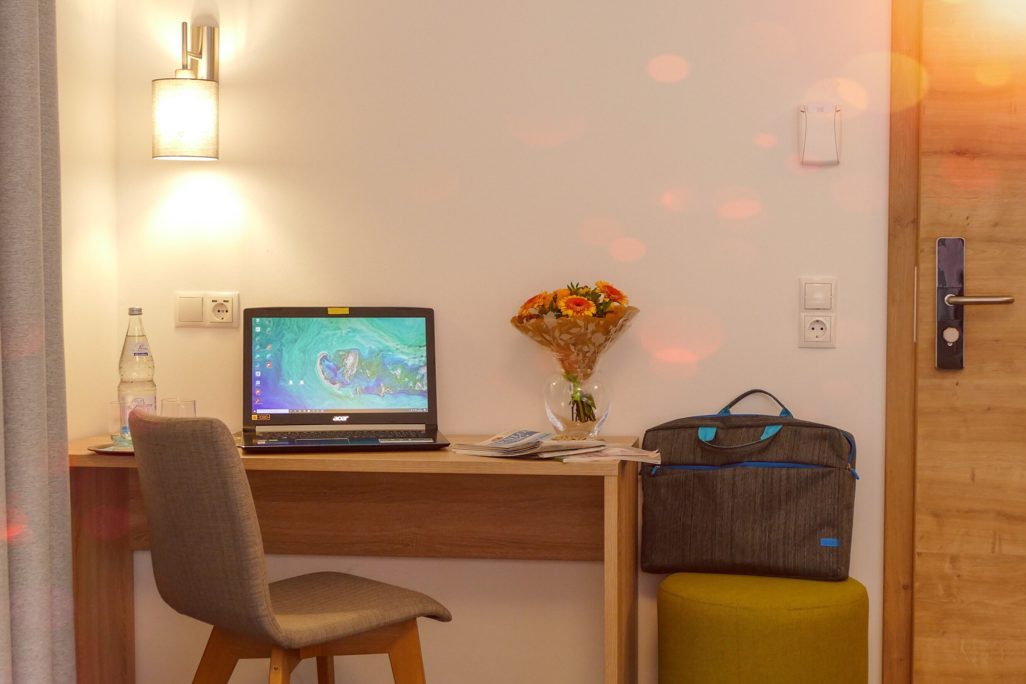 A hotel room with a wooden desk, a laptop, a vase with flowers, a suitcase, and a chair.