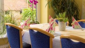 Un comedor con una mesa dispuesta para dos personas, rodeada de sillas tapizadas en azul. Hay plantas en macetas sobre la mesa, y un ventanal ofrece vistas al follaje exterior.