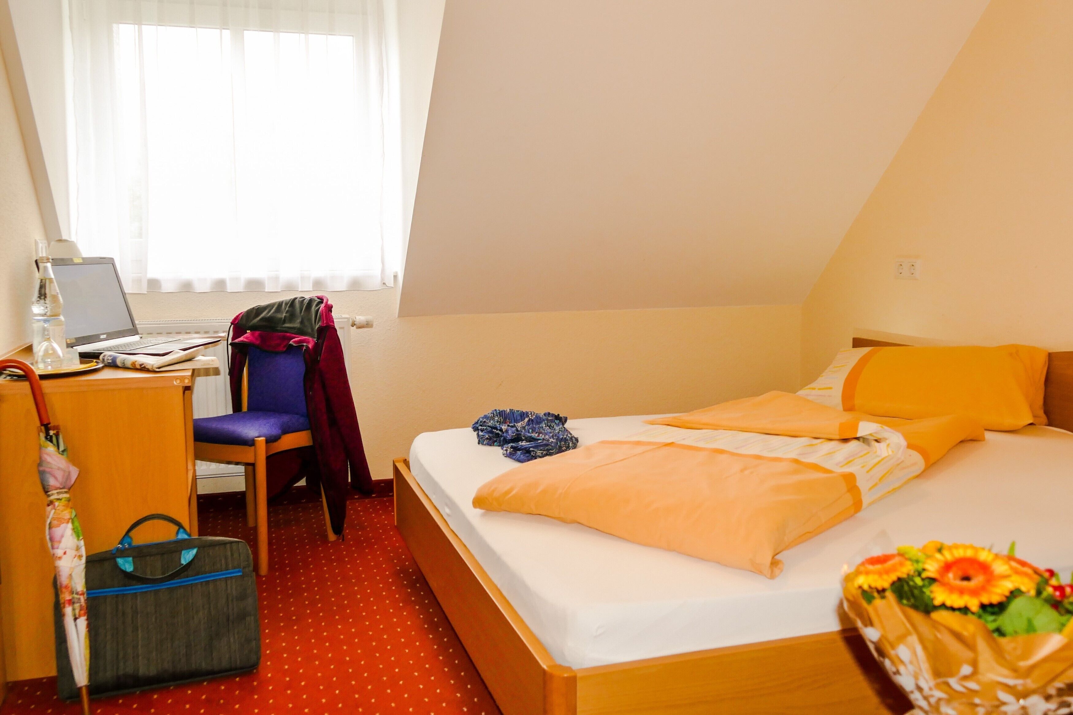 Habitació d'hotel amb un llit, una escriptori amb un ordinador portàtil, una cadira, una maleta i una finestra amb cortines.