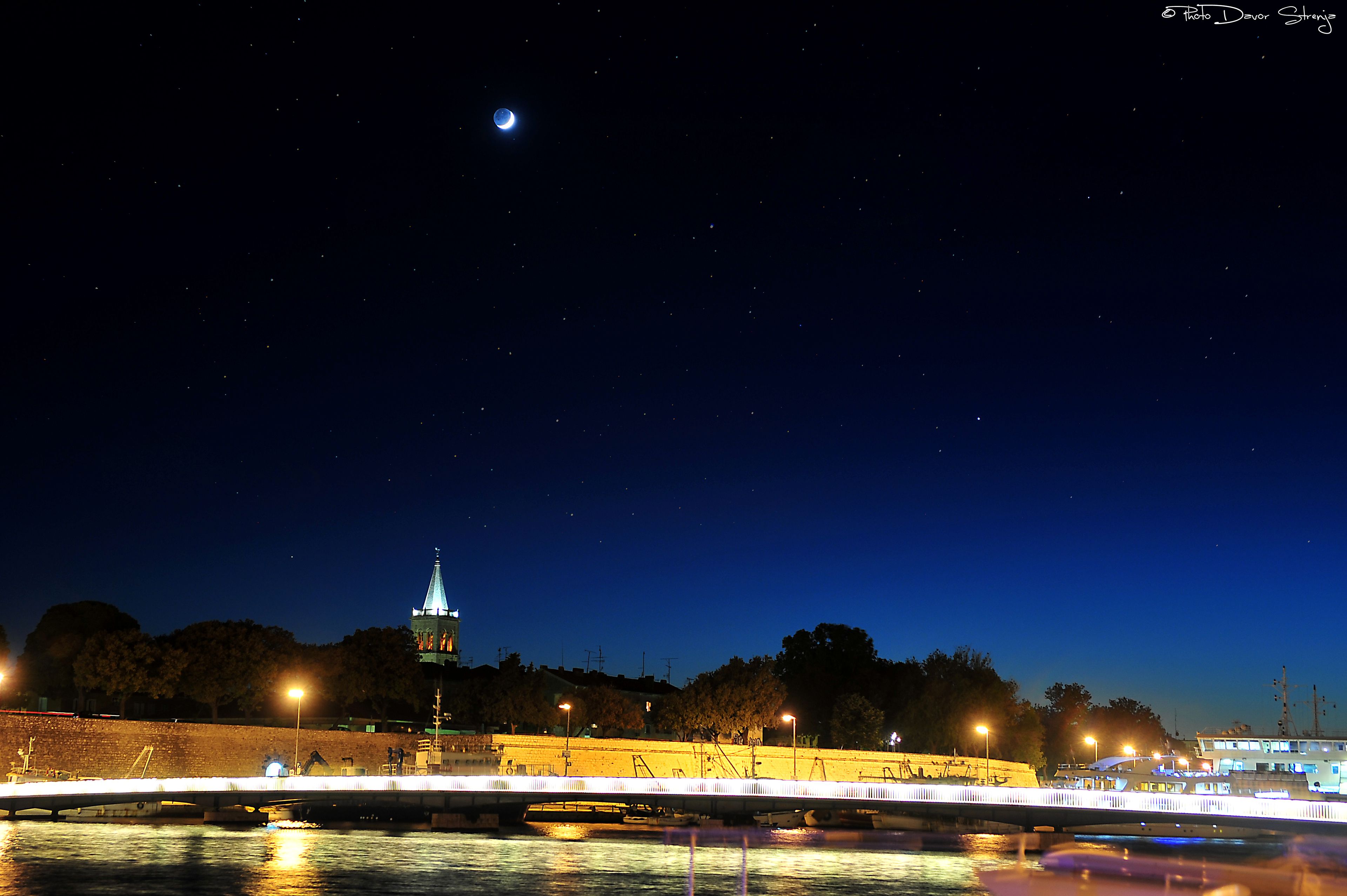 A nighttime cityscape with a bridge, illuminated buildings, and a visible moon.
