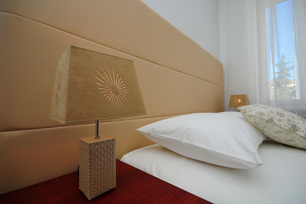 Ein ordentlich bezogenes Bett mit weißen Leinentüchern, eine dekorative Lampe mit geometrischer Basis und ein Fenster mit transparenten Vorhängen.