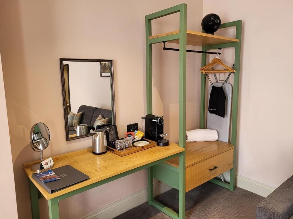 A compact room with a desk, a mirror, a coffee maker, a hanging coat, and a bed.