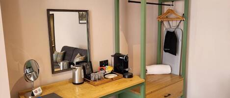 Quarto pequeno com escrivaninha, espelho, cafeteira, cabide e cama.