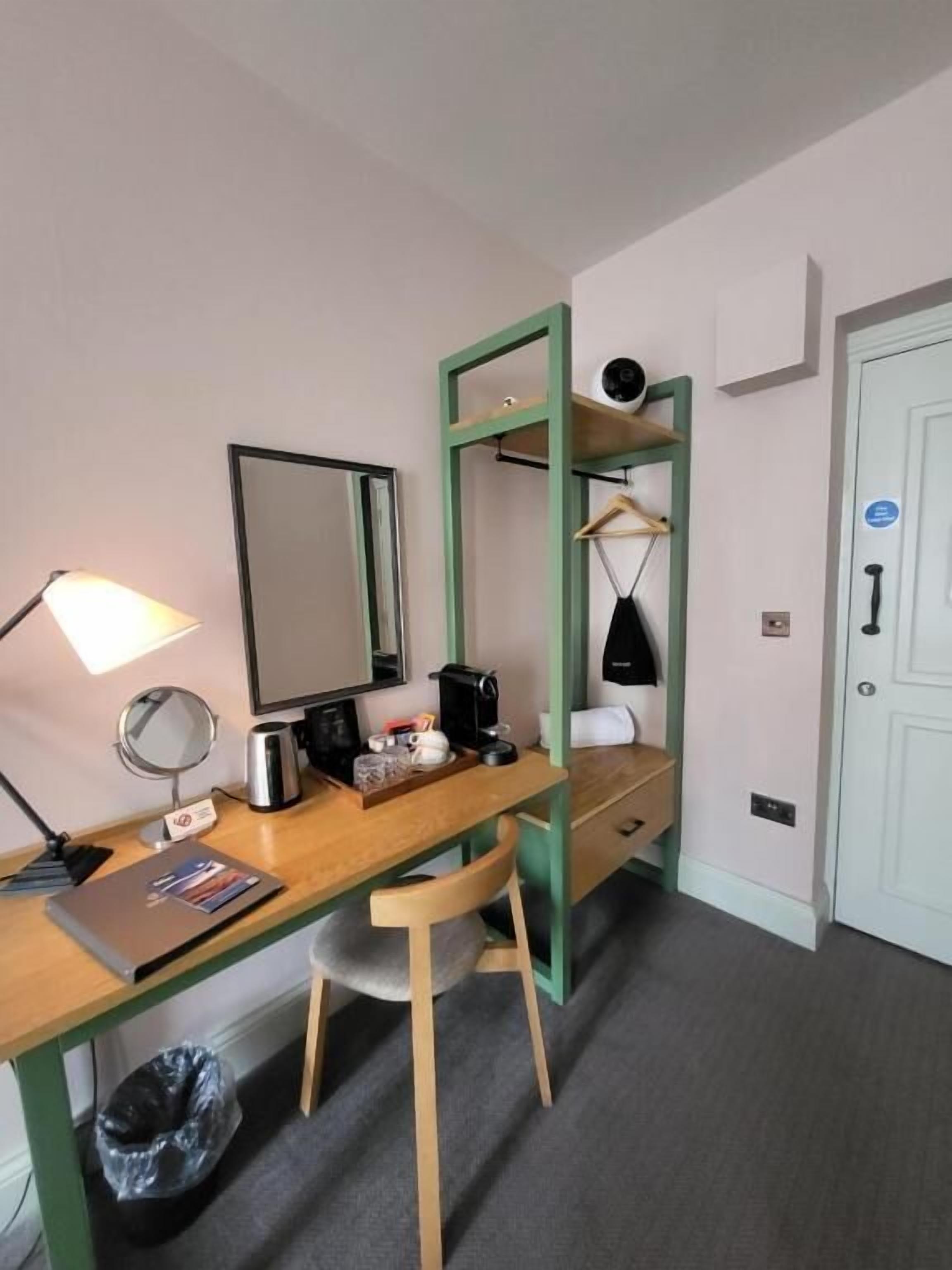 Habitación de hotel compacta con escritorio de madera, espejo, lámpara, cafetera, repisa con bolso negro y puerta blanca.