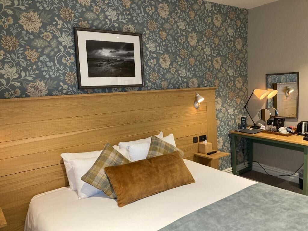 A hotel room with a wooden headboard, a bed with white and brown bedding, a nightstand with a lamp, a desk with a computer and a mirror, and a framed picture on the wall.