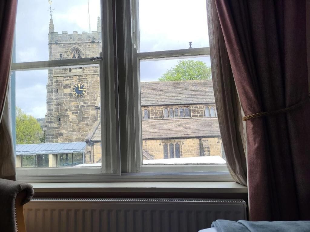 Una ventana con vista a una torre de piedra con un reloj, un radiador debajo y cortinas entreabiertas.
