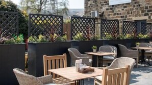 Un patio al aire libre con mobiliario de madera, un edificio de piedra al fondo y una zona delimitada con plantas.