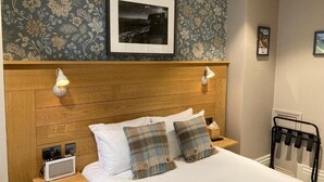 Una habitación de hotel con cabecera de madera, una cama con ropa blanca, dos almohadas a cuadros, una mesita de noche con lámpara, un cuadro enmarcado en la pared y un radiador.