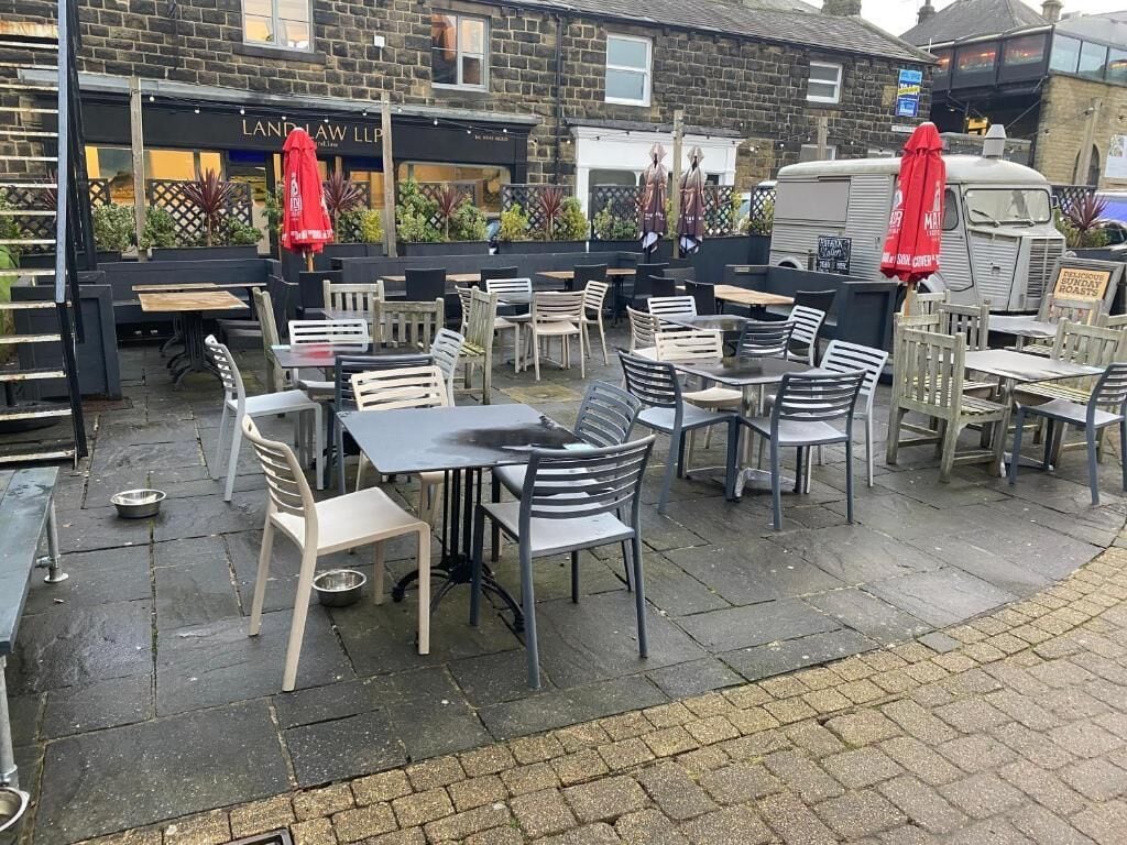An outdoor cafe with tables and chairs, a brick building in the background, and a parked vehicle.