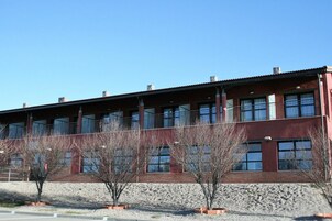 Un edificio de varios pisos con fachada de ladrillo rojo, ventanas grandes y techo plano. Hay árboles desnudos frente al edificio.