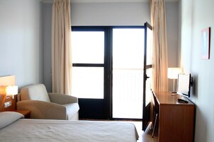 Habitación de hotel con cama, una silla, un escritorio, un televisor y un ventanal grande con cortinas.
