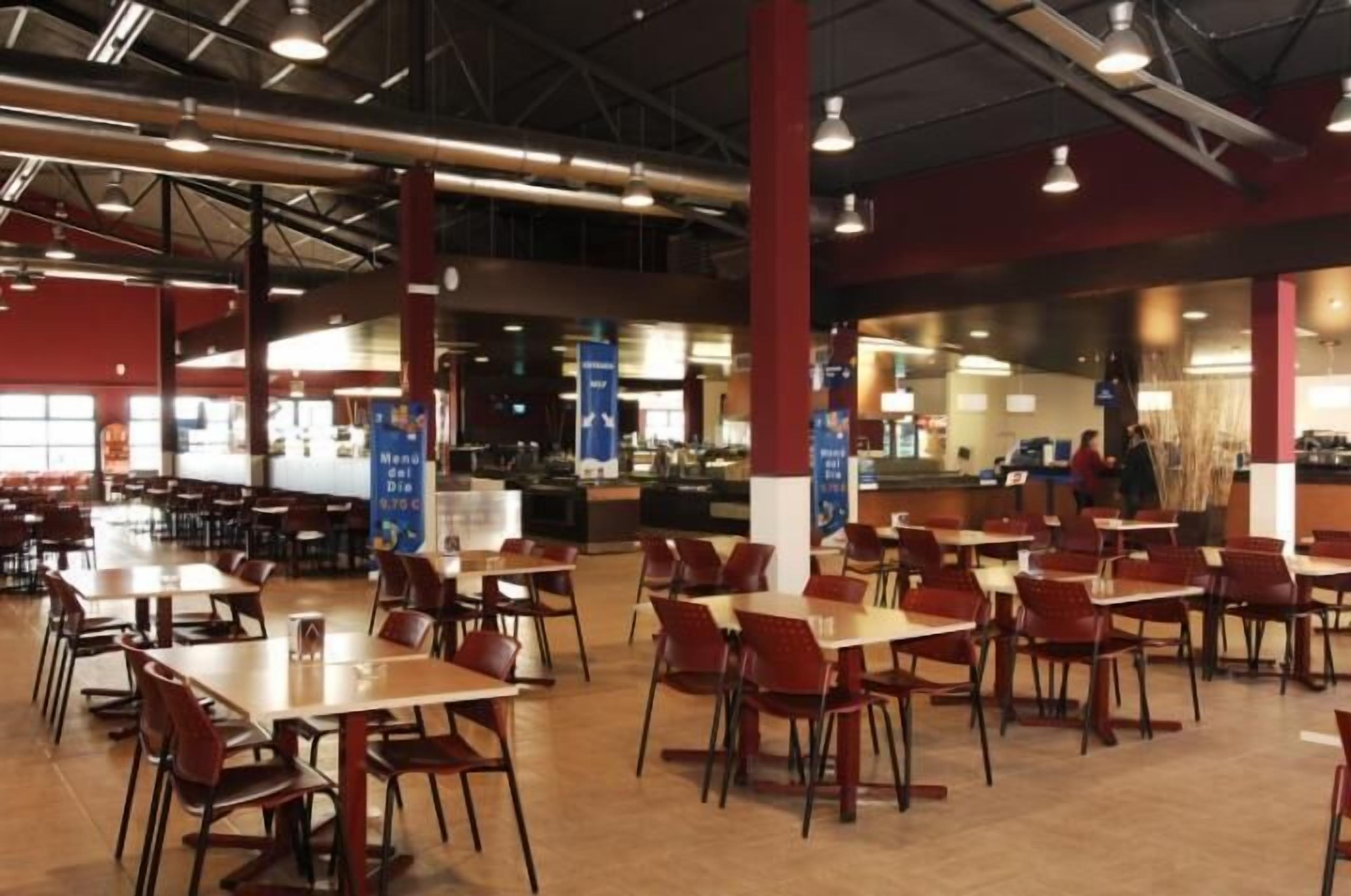 Una amplia cafetería con varias mesas y sillas, una zona de mostrador y un panel de menú visible.