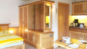 Een kamer met een bed, een houten kledingkast, een tafeltje met een stoel en een kitchenette.