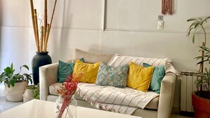 A modern living room with a white sofa, colorful cushions, a white coffee table, and a large vase with dried plants. There is a macrame wall hanging and a potted plant in the corner. - Hotel Sienna (Mendoza)